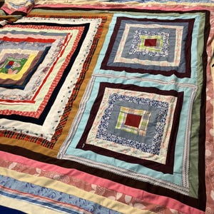 Peut inclure: Une courtepointe en patchwork au motif de cabane en rondins, avec deux grands blocs carrés à droite et une large section rectangulaire à gauche. Elle est composée de bandes de tissu variées dans des tons de bleu, rose, marron, rouge et blanc.