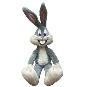 Può includere: Un peluche di Bugs Bunny in posizione seduta. Il peluche è prevalentemente grigio con dettagli bianchi sul petto, sui piedi e sul viso. Le lunghe orecchie sono dritte e la bocca è aperta in un ampio sorriso, rivelando grandi denti. Un personaggio dei cartoni animati classico.