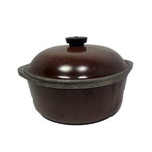 Könnte beinhalten: Ein brauner Kochtopf aus Gusseisen mit passendem Deckel und schwarzem Griff. Der Topf hat einen silbernen Rand. Der Topf ist rund und hat ein klassisches Design.