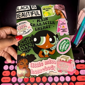 May include: A green sticker sheet with various designs and text, including "BLACK IS BEAUTIFUL," "MAIN CHARACTER ENERGY," and "LET'S GET THIS BREAD." The sheet features cartoon illustrations and a keyboard in the foreground.