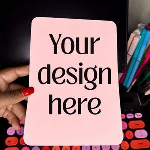 May include: A rectangular light pink sign with rounded corners featuring the black text Your design here. The sign is held in front of a black computer keyboard with colorful keys and a clear container filled with various colored pens and markers.