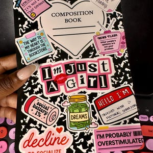 May include: A black and white marble-patterned composition book cover decorated with various colorful stickers. Text includes "I'm Just A Girl," "Reminder: It's Not That Serious," "The way to my heart is through the bookstore," and "I'm probably overstimulated."