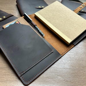 May include: A collection of brown leather notebooks and organizers. One organizer is open, revealing a tan notebook and a pen. The leather has a distressed look, and the stitching is visible. The organizers have a snap closure.