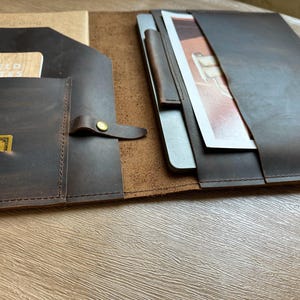 May include: A dark brown leather folio with multiple compartments. One section holds a tablet and a pen, while another features a pocket with a notepad and a credit card. The folio is secured with a leather strap and a brass button.