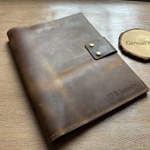 Op de afbeelding: Een bruine leren portfolio met een klepsluiting die is vastgezet met twee messingkleurige studs. De portfolio heeft de initialen "DB Jackson" op de voorkant gegraveerd. Een houten onderzetter met de tekst "GermaFinds" ligt ernaast.