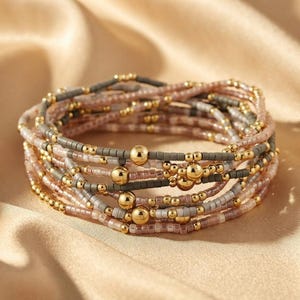 May include: A multi-strand bracelet composed of numerous small beads in shades of dusty rose, muted grey, and metallic gold. The design features alternating rows of tiny seed beads and larger, smooth, polished gold-tone spherical accent beads.