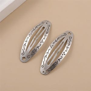 May include: Two oval-shaped silver-toned metal hair clips featuring a cut-out design with engraved swirling patterns and small flower-shaped accents at the ends. The clips have a central elongated opening and a hinged mechanism for securing hair.