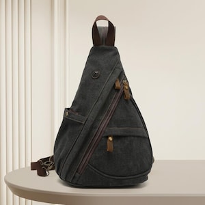 May include: A dark gray canvas sling backpack with a teardrop shape, featuring brown fabric accents, multiple zippered compartments, a top handle, and a small circular logo patch on the upper front section. The bag includes brown leather zipper pulls.