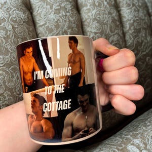 May include: A white ceramic mug with a collage of shirtless men and the text "I'M COMING TO THE COTTAGE". The mug is held by a person with pink nail polish.