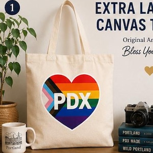May include: An off-white canvas tote bag featuring a large heart-shaped graphic on the front. The heart displays the Progress Pride flag colors with the white text PDX centered inside. The bag has two long shoulder straps and a rectangular shape.