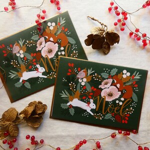 May include: Two Christmas cards with a green background and gold accents. The cards feature a white rabbit, a squirrel, a robin, and flowers. The text "Noel" is written in gold on each card.
