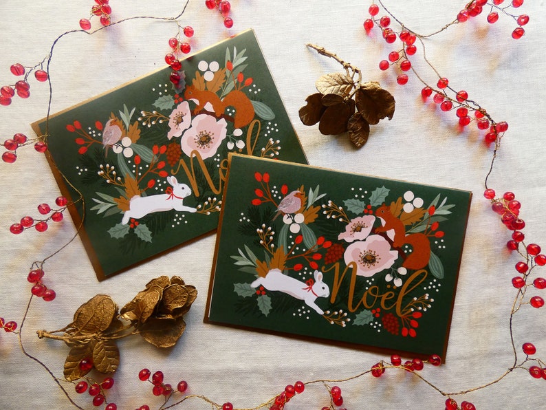 May include: Two Christmas cards with a green background and gold accents. The cards feature a white rabbit, a squirrel, a robin, and flowers. The text "Noel" is written in gold on each card.