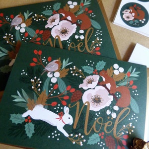May include: Two green Christmas cards with a festive woodland scene. The cards feature a white rabbit, red squirrels, and red berries on a green background. The text "No&euml;l" is written in gold on both cards.