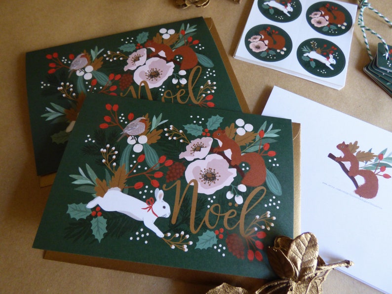 May include: A set of Christmas cards with a green background and gold accents. The cards feature a festive design with a white rabbit, red squirrels, and a floral pattern. The text "No&euml;l" is written in gold script.