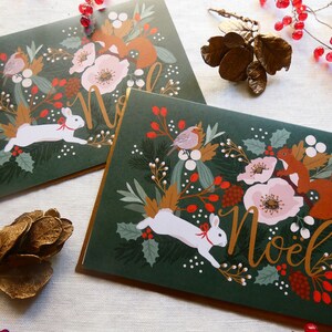 May include: Two green Christmas cards with gold lettering that says "Noel". Each card features a white rabbit, a squirrel, and a robin surrounded by red berries, flowers, and greenery.