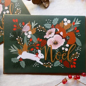 May include: A green Christmas card with a gold border features a white rabbit, a squirrel, and a robin surrounded by flowers, berries, and leaves. The card says "No&euml;l" in gold lettering.