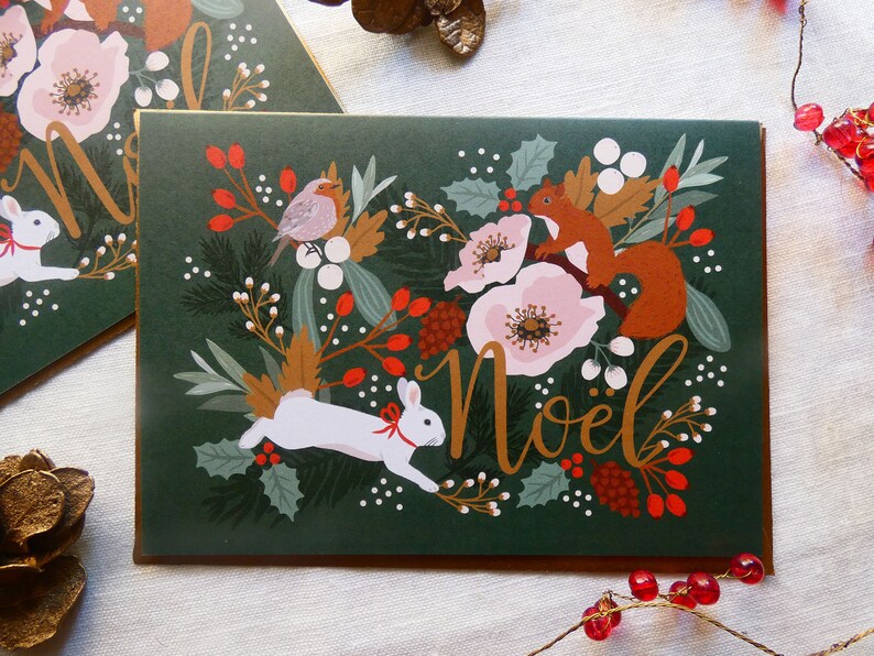 May include: A green Christmas card with a gold border features a white rabbit, a squirrel, and a robin surrounded by flowers, berries, and leaves. The card says "No&euml;l" in gold lettering.