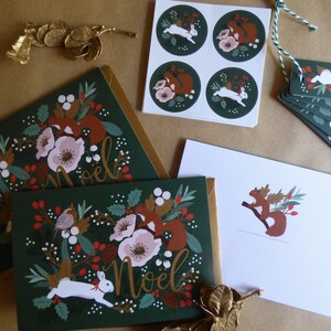 May include: A set of Christmas cards and gift tags featuring a green background with white rabbits, red squirrels, and red berries. The cards and tags have a gold foil accent and the text "Noel" in gold lettering.