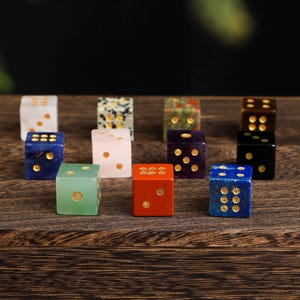 May include: A collection of twelve gemstone dice in various colors, including white, blue, green, and red. Each die features gold-colored dots. The dice are arranged on a dark wooden surface, showcasing their unique textures and hues.