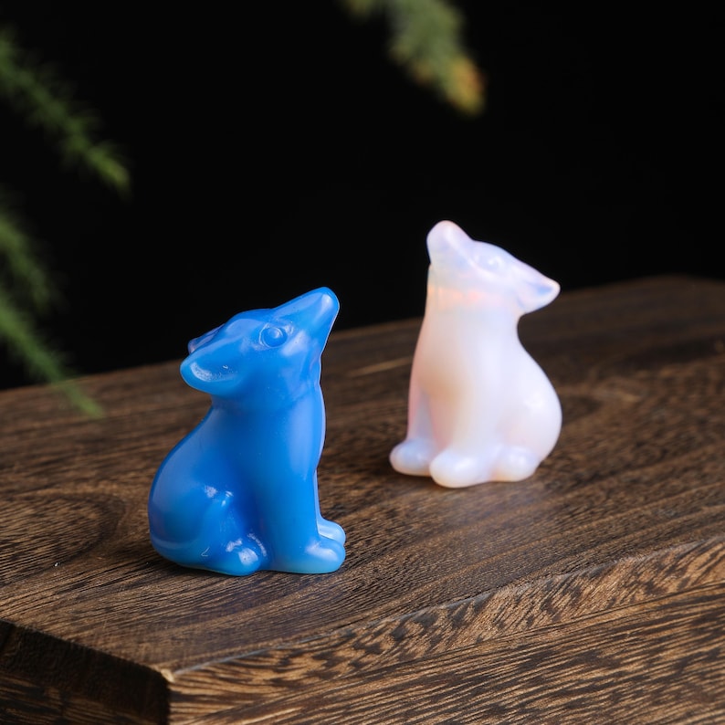 May include: Two small, carved wolf figurines. One is a vibrant blue, the other is a translucent white. Both are posed with their heads tilted upwards. They are displayed on a dark wooden surface.