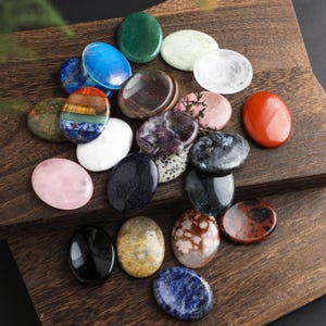 Può includere: Una collezione di pietre preziose ovali e lucide in vari colori, tra cui blu, verde, rosa e rosso. Le pietre sono disposte su una superficie di legno scuro, mettendo in risalto le loro trame e motivi naturali. Le pietre preziose variano per dimensioni e opacità.