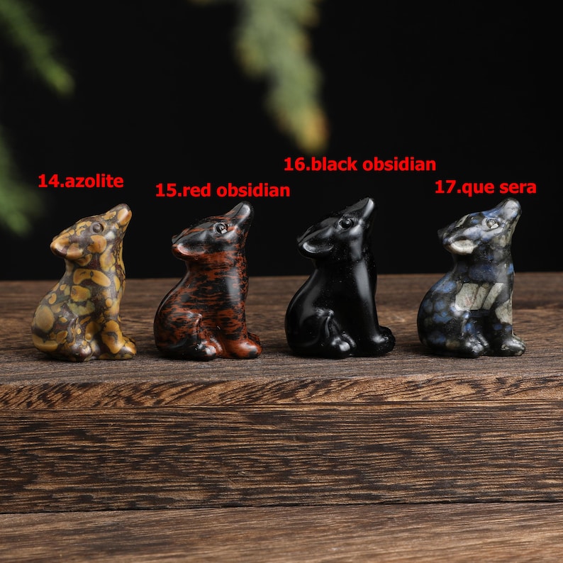 May include: Four small, carved stone wolf figurines in a row. The azolite wolf is tan and brown, the red obsidian wolf is dark red, the black obsidian wolf is solid black, and the que sera wolf is blue and gray. Each wolf is looking up.