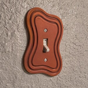 Op de afbeelding: Een terracotta-kleurige lichtschakelaarplaat met een golvende, organische vorm en concentrische gelaagde details. De plaat is voorzien van een centrale tuimelschakelaar in een lichtbeige kleur en wordt bevestigd met twee verticaal geplaatste schroeven.