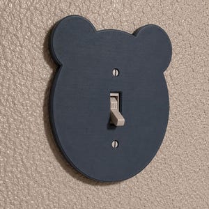 May include: A dark blue, bear-shaped light switch cover plate with two silver screws securing it to the wall. The plate features a central rectangular cutout for a beige toggle switch, which is currently in the downward position labeled OFF.