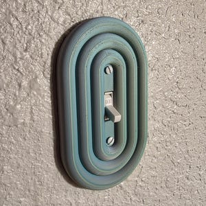 Könnte beinhalten: Eine hellblaue, 3D-gedruckte Lichtschalterabdeckung mit ovalem Design. Die Abdeckung hat ein geschichtetes, konzentrisches Muster um den Schalter. Der Schalter selbst ist weiß, und die Abdeckung ist an einer strukturierten, hellgrauen Wand befestigt.