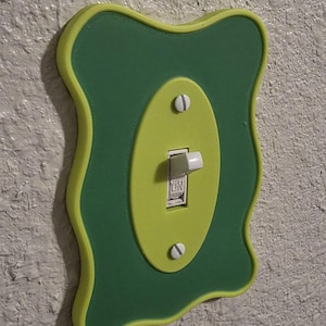 Pode incluir: Uma capa decorativa para interruptor de luz com uma borda ondulada e irregular. A capa é principalmente verde com um centro ovalado verde mais claro. O interruptor em si é branco e os parafusos são prateados. O interruptor está na posição "ON".