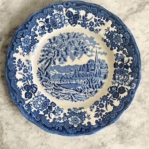Peut inclure: Une assiette décorative ronde bleue et blanche avec une bordure florale. Le centre présente un paysage avec un arbre, des bâtiments et une église. L'assiette a un bord festonné.