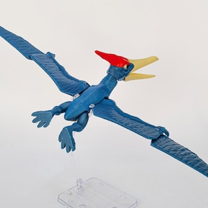 Op de afbeelding: Een blauwe pterosauriër dinosaurus actiefiguur met een rode kam en een lichtgele snavel. Het figuur is afgebeeld met de vleugels volledig gespreid en wordt ondersteund door een doorzichtige plastic standaard onder het lichaam.