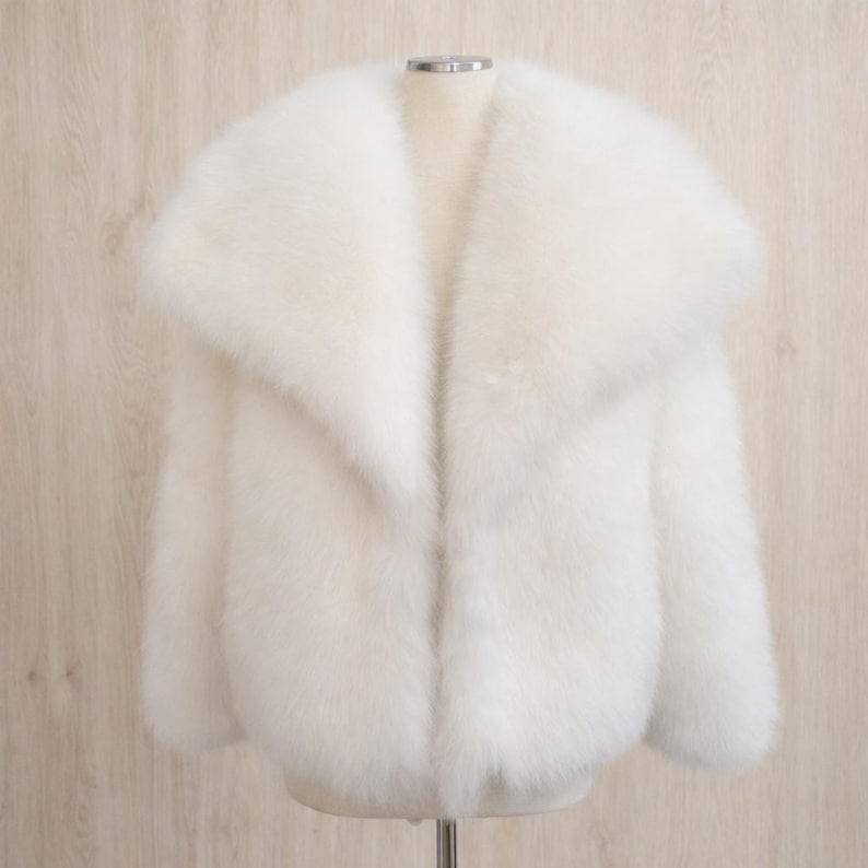 May include: A fluffy, white faux fur jacket with a wide collar. The jacket is displayed on a mannequin, showcasing its soft texture and voluminous design. The sleeves are long, and the jacket appears to be a cropped style.
