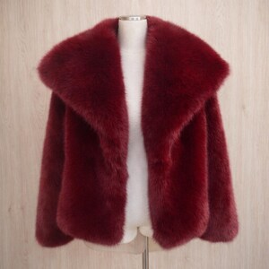 May include: A burgundy faux fur jacket with a wide collar. The jacket is displayed on a mannequin against a light wood-grain background. The jacket has long sleeves and a soft, plush texture.