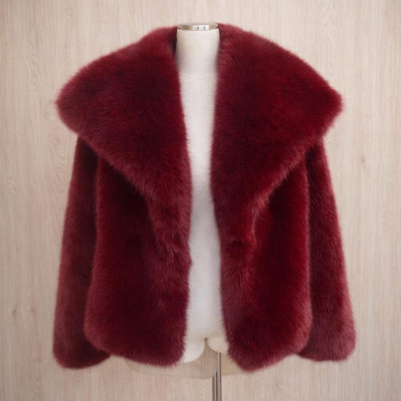 May include: A burgundy faux fur jacket with a wide collar. The jacket is displayed on a mannequin against a light wood-grain background. The jacket has long sleeves and a soft, plush texture.