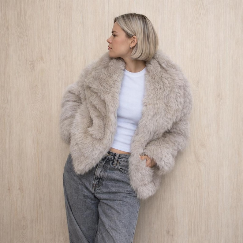 May include: A light beige faux fur coat with a wide collar. The coat is worn over a white t-shirt and paired with gray jeans. The model is standing in front of a light wood-grain background.