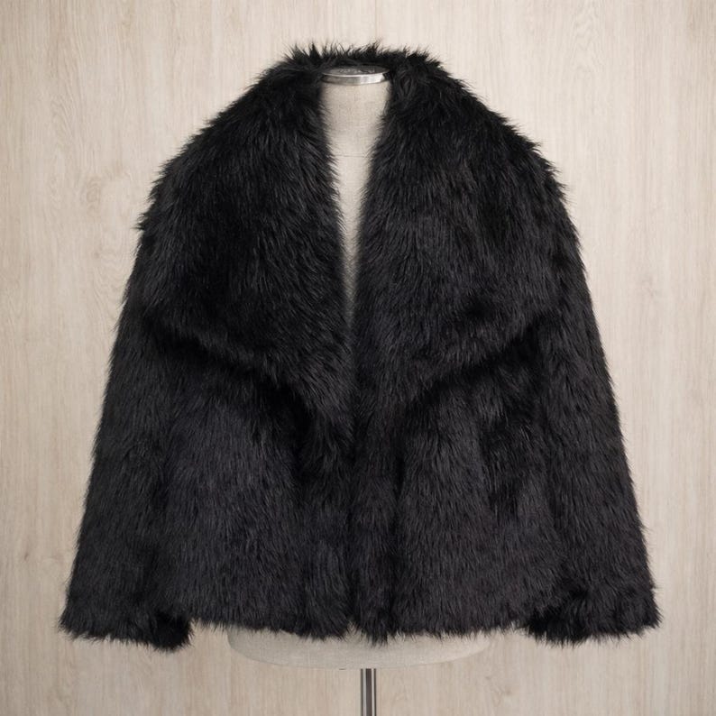 May include: A black faux fur jacket with a wide collar. The jacket is displayed on a mannequin against a light wood-grain background. The jacket has a soft, textured appearance.
