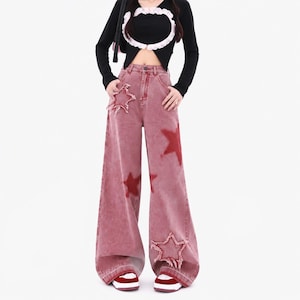 Può includere: Jeans a gamba larga in denim rosso slavato con vita alta, cinque tasche e chiusura a bottoni. I pantaloni sono decorati con diverse stampe di stelle rosse e due vistose applicazioni a forma di stella con bordi sfrangiati sulle gambe.