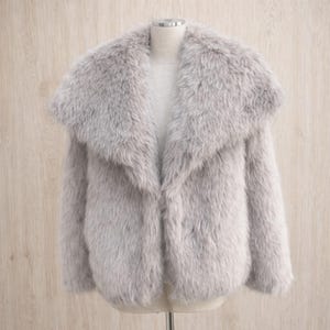 May include: A light gray faux fur coat with a wide collar. The coat is displayed on a mannequin against a light wood background. The coat has long sleeves and a soft, textured appearance.