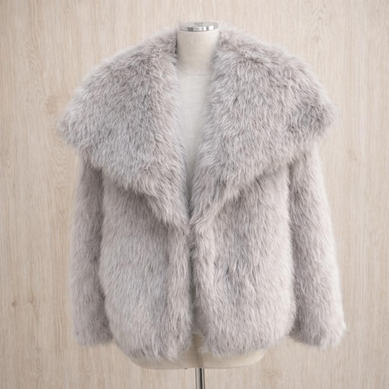 May include: A light gray faux fur coat with a wide collar. The coat is displayed on a mannequin against a light wood background. The coat has long sleeves and a soft, textured appearance.