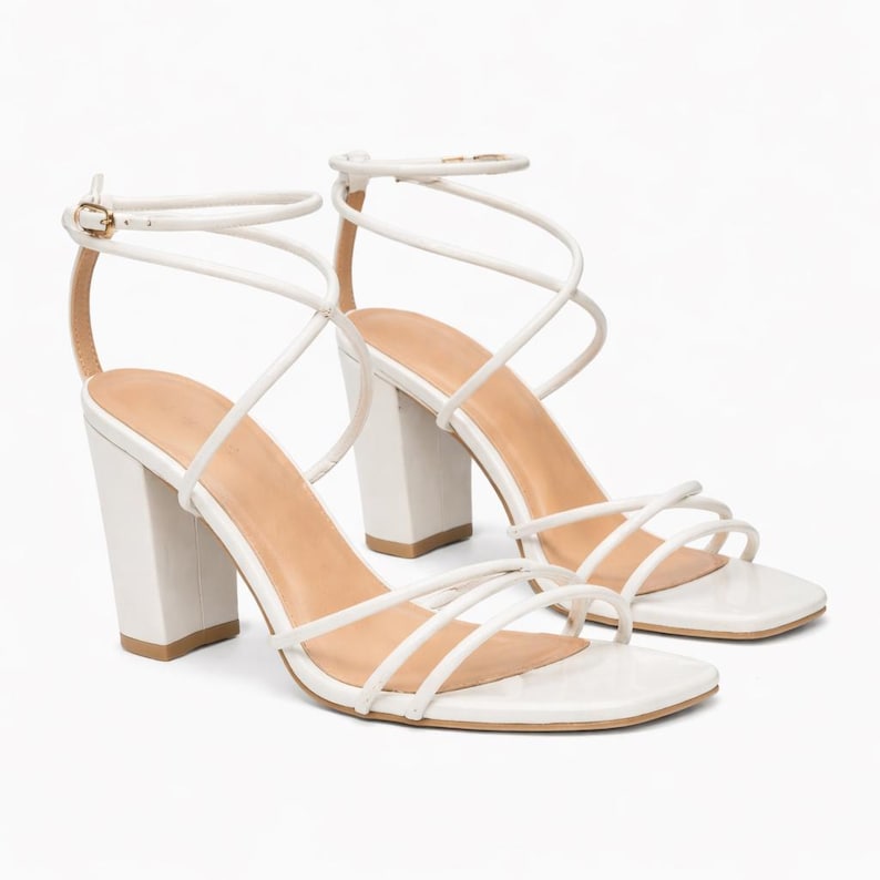 May include: A pair of white high-heeled sandals with a square toe and multiple thin straps. The sandals have a block heel and an ankle strap with a gold-colored buckle. The footbed is a light tan color.