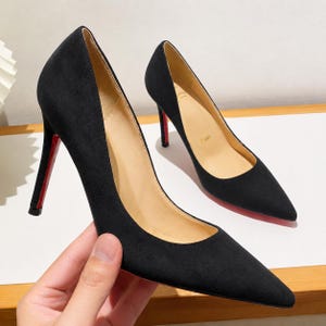May include: A pair of black suede pumps with a pointed toe and a high stiletto heel. The interior lining is beige, and the heel features a distinct red accent on the back. The word Paris is embossed on the beige insole of the shoe.