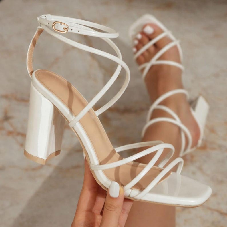 May include: White strappy heeled sandals with a square toe and a thick block heel. The ankle strap features a gold-colored buckle. The sandals are shown on a light-colored surface.