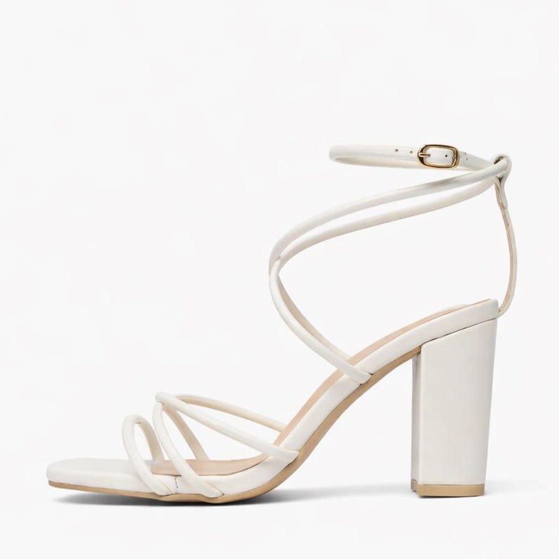 May include: White strappy heeled sandal with a square toe and ankle strap. The sandal features multiple thin straps that wrap around the foot and ankle. The block heel is approximately 3.5 inches high.