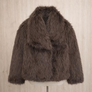 May include: A brown faux fur jacket with a wide collar. The jacket has a button closure and a soft, textured appearance. The garment is displayed on a mannequin against a light wood background.