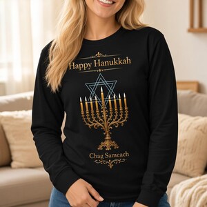 May include: A black long-sleeve sweatshirt featuring a gold-colored menorah with lit candles, a blue Star of David outline, and the text "Happy Hanukkah" at the top and "Chag Sameach" at the bottom, paired with blue denim jeans.