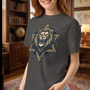 May include: A charcoal gray short-sleeve t-shirt featuring a graphic print of a roaring lion head centered within a gold and dark blue six-pointed star emblem. The design is positioned on the chest of the garment, which is paired with light blue jeans.