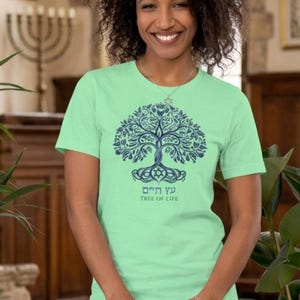 May include: A light green short-sleeved t-shirt featuring a dark blue graphic of a tree with intricate branches and roots. Below the tree, the Hebrew text "עץ חיים" is printed above the English text "TREE OF LIFE" in a dark blue font.