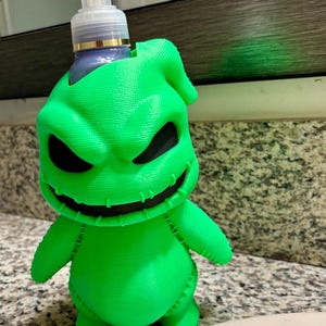 Peut inclure: Un porte-savon liquide vert n&eacute;on vif en forme de personnage avec un large sourire cousu et des yeux sombres et creux. Le support pr&eacute;sente une surface textur&eacute;e imprim&eacute;e en 3D et contient un flacon pompe en plastique transparent avec un embout blanc.