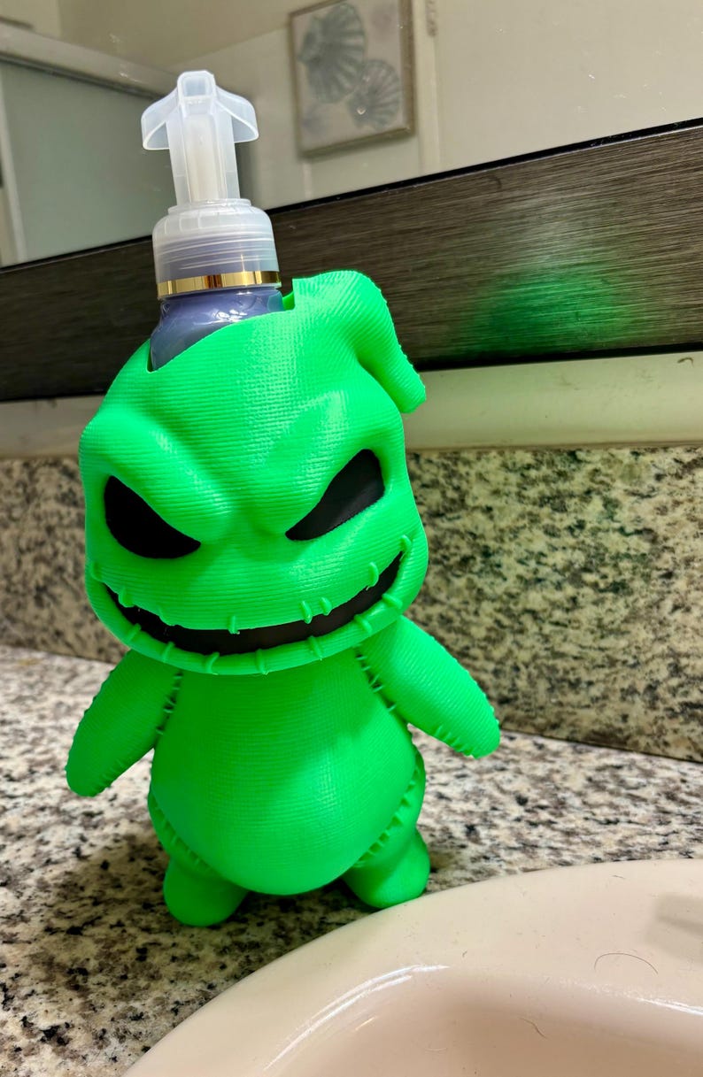 Peut inclure: Un porte-savon liquide vert n&eacute;on vif en forme de personnage avec un large sourire cousu et des yeux sombres et creux. Le support pr&eacute;sente une surface textur&eacute;e imprim&eacute;e en 3D et contient un flacon pompe en plastique transparent avec un embout blanc.